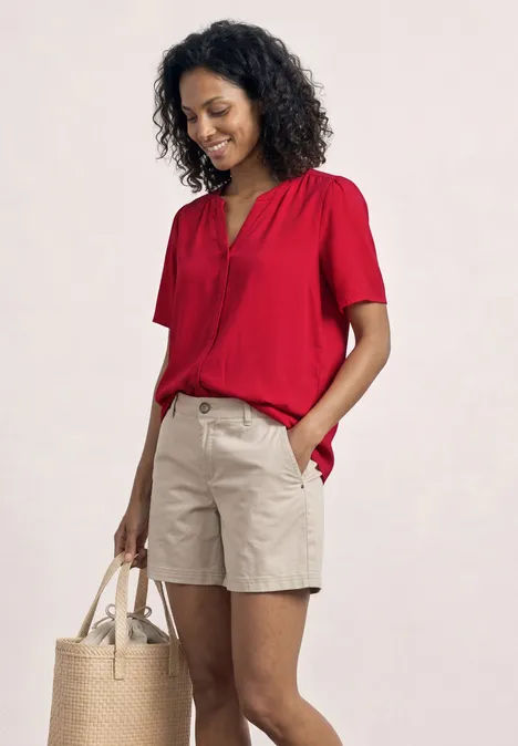 Une personne porte un top rouge à manches courtes, un short beige et tient un sac en paille.