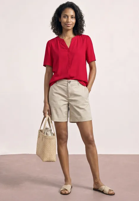 Une personne en chemisier rouge, short beige et sandales porte un sac en paille sur fond clair.