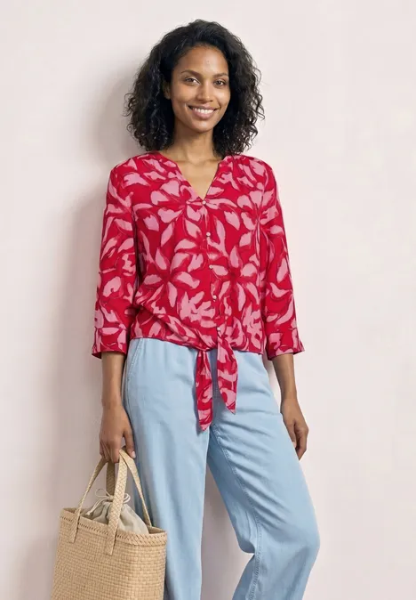 Frau trägt rote Blumenbluse mit Knoten, hellblaue Hose und handliche Tasche.
