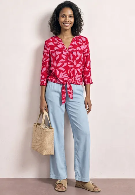 Frau trägt rote, gemusterte Bluse, vorne geknotet, weite hellblaue Jeans, Sandalen und Strohtasche.