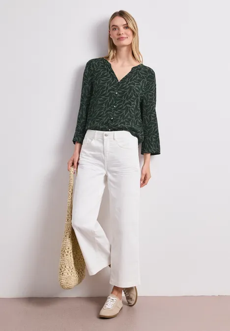 Frau in dunkelgrüner Bluse, weißer Culotte, goldenen Schuhen hält Korbtasche vor hellem Hintergrund.