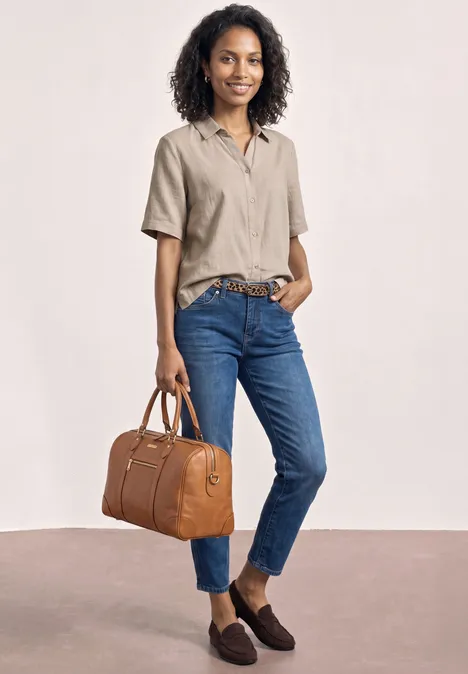 Frau in beigefarbenem Hemd und blauen Jeans mit braunen Loafers hält eine braune Lederhandtasche.