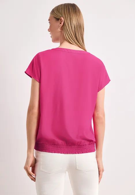 Vrouw van achter gefotografeerd in een korte fuchsia roze top met witte broek tegen witte achtergrond.