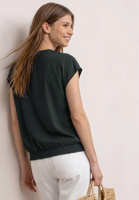 Vrouw van achteren in zwart shirt, witte broek en strozak, staand voor een lichte muur.