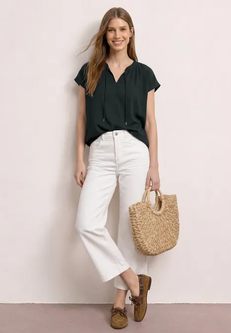 Vrouw in zwart shirt en witte jeans, met bruine loafers en strooien tas tegen een lichte achtergrond.