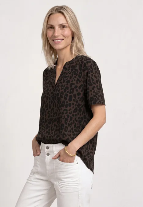 Frau mit kurzärmliger Bluse im Leopardenmuster und weißer Jeans, Hände in den Taschen.