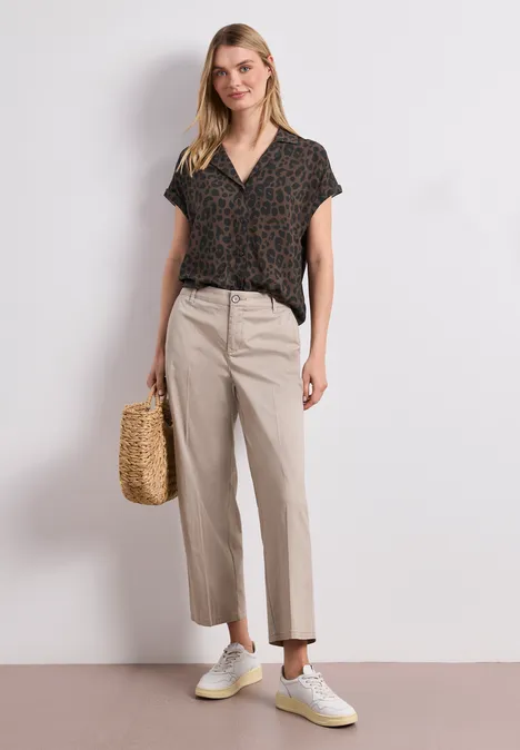 Femme en haut à motif, pantalon beige, baskets blanches et sac en paille sur fond neutre.
