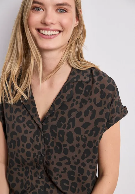 Jeune femme aux cheveux blonds porte une chemise brun foncé à motif léopard noir et sourit.