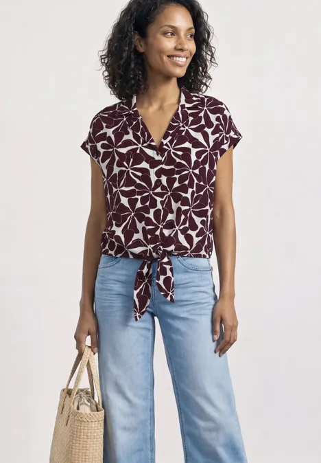 Frau mit gemustertem Kurzarm-Top in Bordeaux und Hellblauem Jeans, Tasche in der Hand.