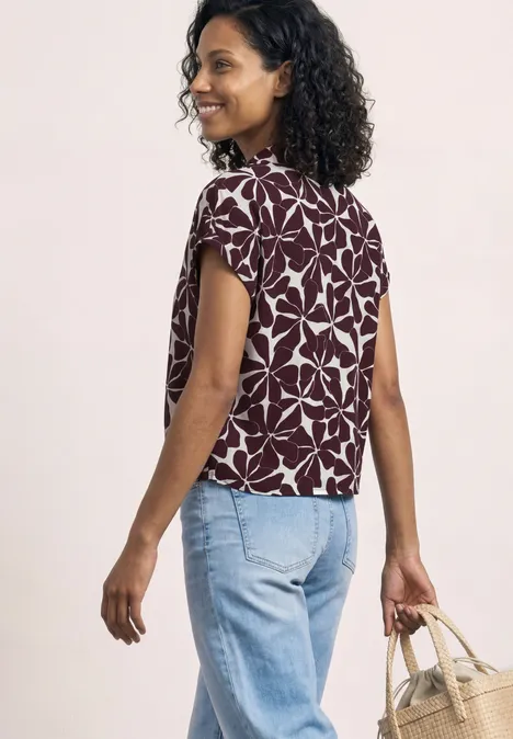 Frau von hinten mit lockigem Haar, floral gemustertem Shirt, hellen Jeans und geflochtener Handtasche.