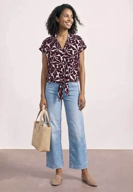 Person trägt violettes Blatt-Print-Top, weite Jeans, Basttasche und braune Schuhe.