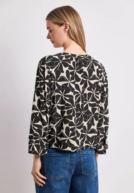 Frau von hinten in schwarz-weiß gemusterter Bluse und blauer Jeans, Kopf leicht zur Seite gedreht.