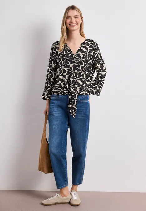 Frau trägt schwarze, gemusterte Bluse mit Knoten, blaue Jeans, helle Sneaker und eine braune Tasche.