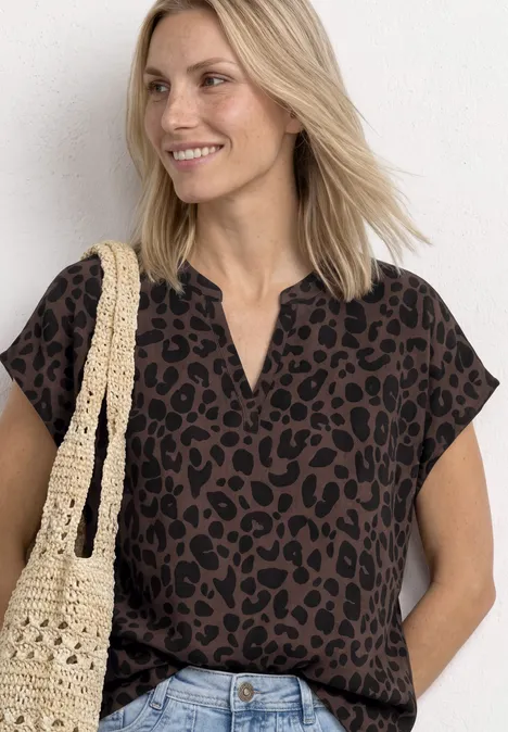 Frau im Leopardenmuster-Top, trägt Jeans und gehäkelte Tasche, lockere Haltung vor weißer Wand.