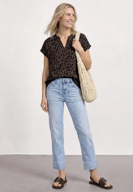 Frau in hellblauen Jeans, gemusterter schwarzer Bluse, Sandalen und Korbtasche.