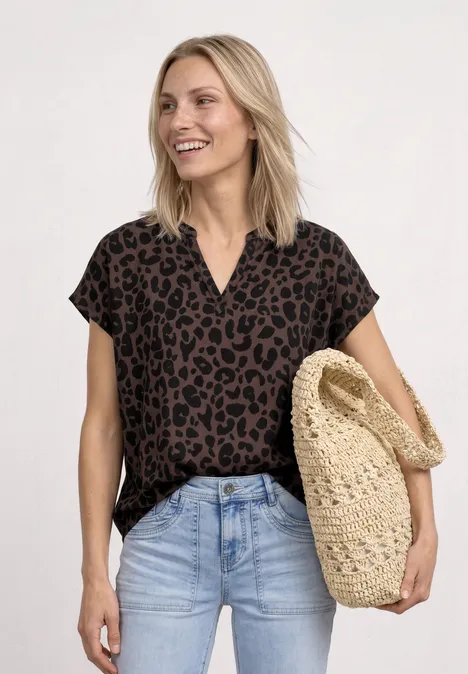 Frau trägt braune Leo-Bluse, helle Jeans und hält eine geflochtene Strohtasche vor hellem Hintergrund.