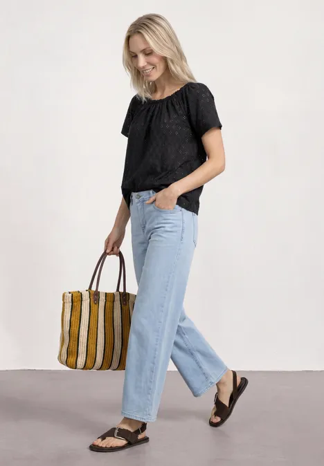 Frau mit schwarzer Bluse, hellblauen Jeans und Sandalen trägt gestreifte Tasche.