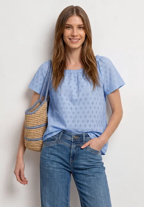 Vrouw draagt lichtblauw kort mouwshirt met patroon, spijkerbroek en rieten schoudertas.