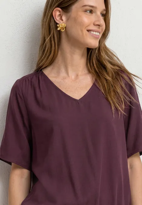 Frau mit langem braunem Haar trägt violette V-Ausschnitt-Bluse und goldene Blumenohrringe vor weißem Hintergrund.