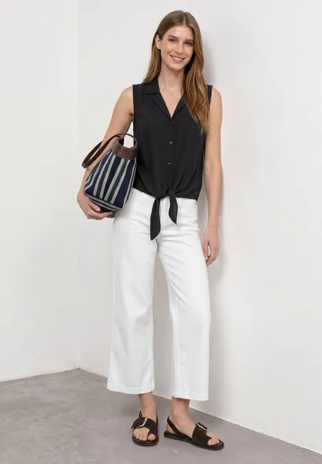 Frau trägt schwarzes ärmelloses Top mit Knotendetail, weiße Culottes, schwarze Sandalen und gestreifte Tasche.