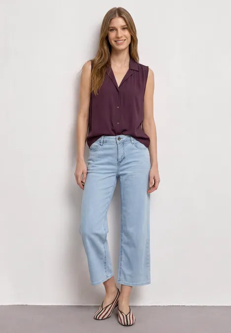 Femme en chemise sans manches violette, jean large bleu clair et sandales devant un mur blanc.