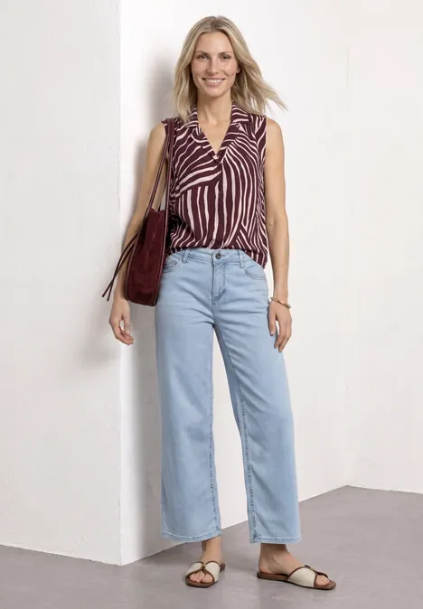 Vrouw voor witte muur, draagt gestreepte mouwloze top, lichte jeans, sandalen en een bordeaux schoudertas.