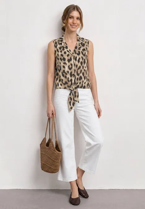 Frau in ärmelloser Leoparden-Print-Bluse, geknotet vorne, weiße Cropped-Hosen, braune Pumps und Korbtasche.