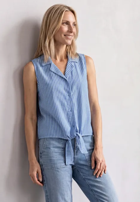 Frau in ärmelloser, blau-weiß gestreifter Bluse mit Knoten vorne und Jeans lehnt an heller Wand.