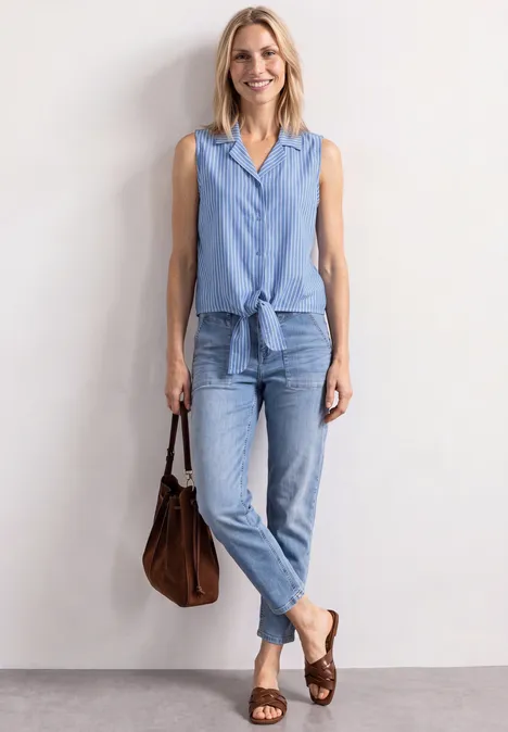 Frau in lässigem, hellblauem Denim-Outfit mit ärmellosem Hemd, Jeans, brauner Ledertasche und Sandalen.