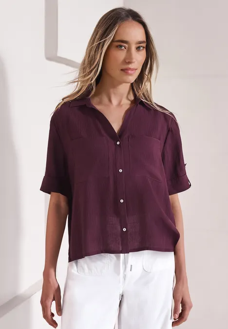 Overhemd met korte mouwen en omgeslagen manchetten rustic rouge Vrouw in een losse paarse blouse met korte mouwen en een witte broek tegen een lichte achtergrond.