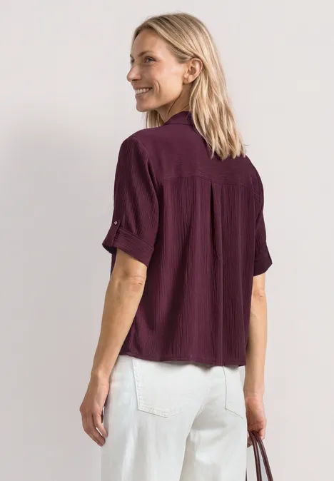 Overhemd met korte mouwen en omgeslagen manchetten rustic rouge Vrouw van achteren met een paarse blouse, witte spijkerbroek en een tas in haar linkerhand.