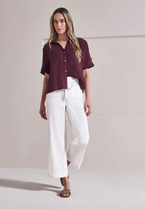 Overhemd met korte mouwen en omgeslagen manchetten rustic rouge Vrouw tegen lichte achtergrond, bordeaux blouse, witte wijde broek en sandalen.