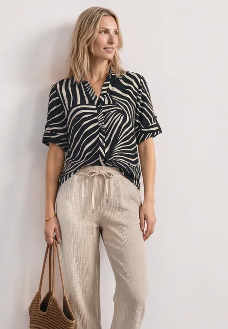 Kurzarm Hemdbluse mit Turn-Up-Details Black Frau in gestreiftem Schwarz-Weiß-Top, beigen Leinenhosen und brauner Tasche.