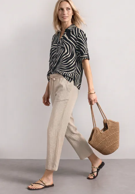 Kurzarm Hemdbluse mit Turn-Up-Details Black Person trägt ein schwarz-weißes Zebra-Muster-Hemd, beige Hose, schwarze Sandalen und hält eine Strohtasche.