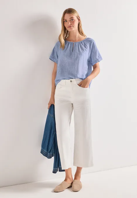 Frau in hellblauer Bluse und weißen Culottes, hält eine Jeansjacke, trägt bequeme Slipper.