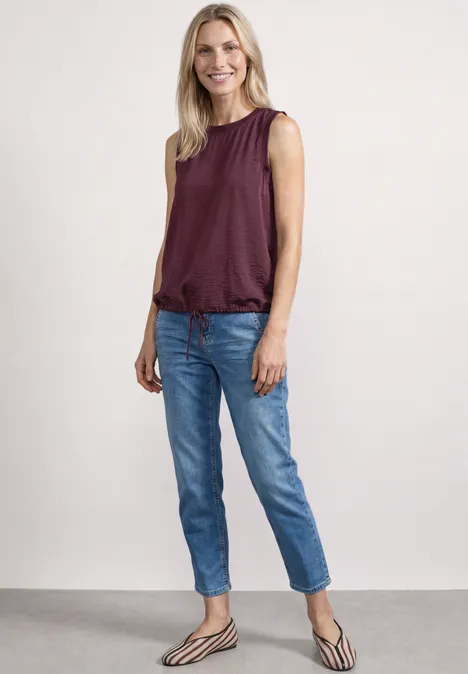 Eine Frau in ärmellosem bordeauxrotem Top, blauen Jeans und gestreiften Ballerinas steht vor hellem Hintergrund.