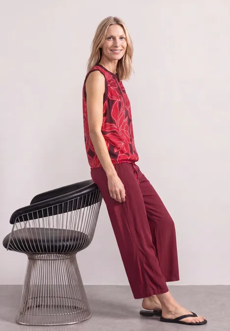 Blusentop mit Rundhals und Tape-Detail salsa red Frau lehnt lässig an einem modernen Stuhl, trägt rotes ärmelloses Oberteil und weite rote Hose.