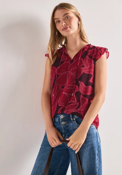 Eine Frau steht vor hellem Hintergrund, trägt eine rote, gemusterte Bluse mit Rüschenärmeln und blaue Jeans.