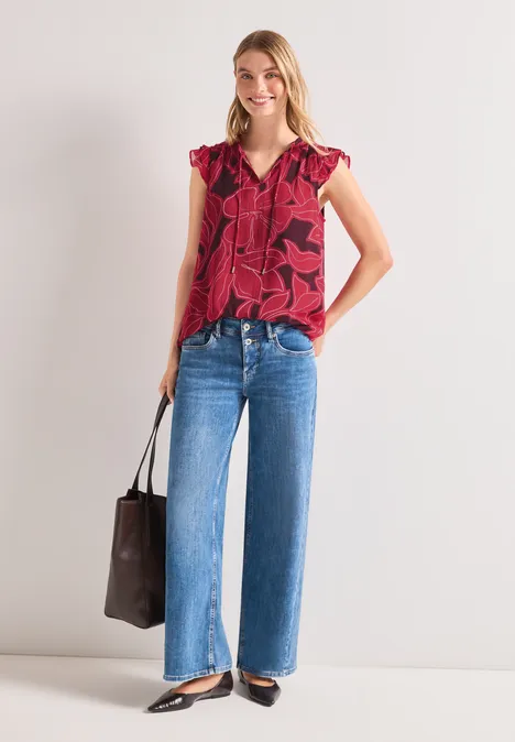 Modell trägt rote, gemusterte Bluse, weite Jeans und schwarze Schuhe, hält braune Tasche vor weißem Hintergrund.