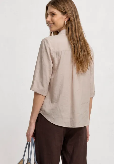 Frau von hinten in einer beigen Hemdbluse und dunklen Hosen, Handtasche in der Hand.