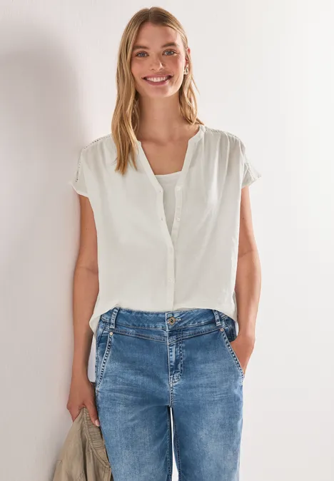 Frau mit weißer Bluse und Jeans, steht vor heller Wand mit Händen in den Hosentaschen.