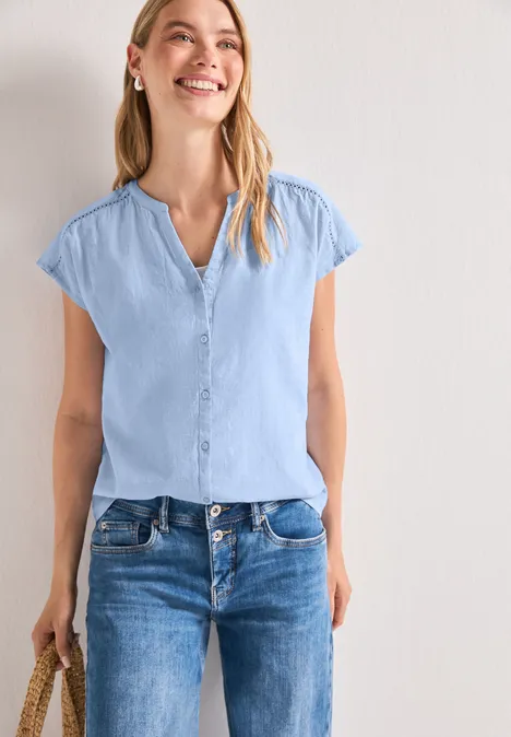 Frau in hellblauer Kurzarmbluse und Jeans hält Strohtasche vor weißer Wand.