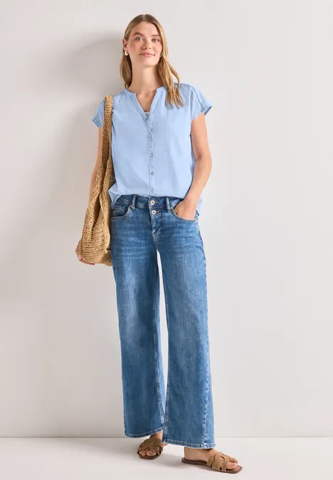 Eine Frau trägt ein hellblaues T-Shirt, weite blaue Jeans, Sandalen und eine Korbtasche vor weißem Hintergrund.