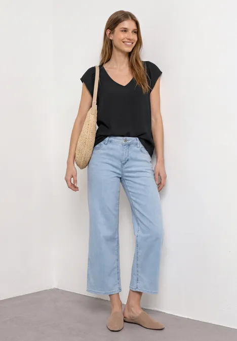 Frau steht in Zimmerecke an weißer Wand, trägt schwarzes V-Shirt, weite hellblaue Jeans, beige Schuhe und Strohtasche.