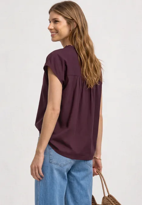 Frau von hinten in dunkelrotem Top und blauer Jeans mit Tasche in der Hand, vor neutralem Hintergrund.