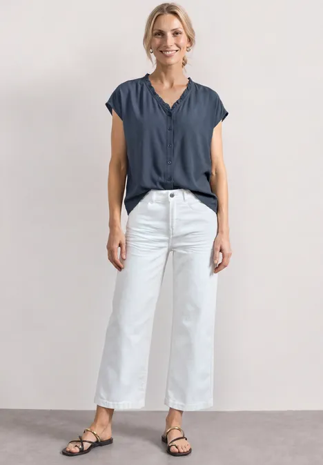 Frau in blauem Oberteil, weißen Culotte-Hosen und schwarzen Sandalen vor neutralem Hintergrund.