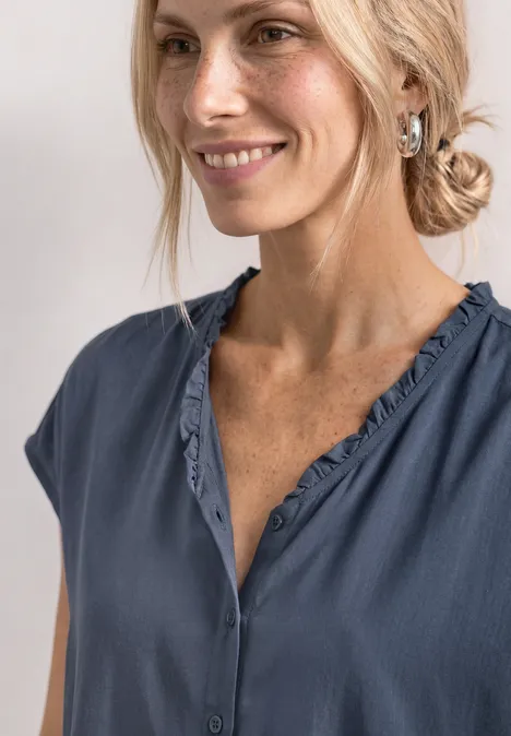 Blonde Frau in blauem, lockerem Oberteil mit V-Ausschnitt, lächelt seitlich in die Kamera.