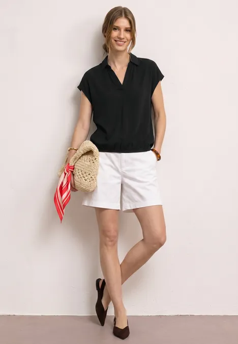 Chemisier à col chemise et ourlet élastiqué Black Femme debout devant un mur blanc, en blouse noire, jupe blanche, ballerines noires et panier avec foulard rouge.