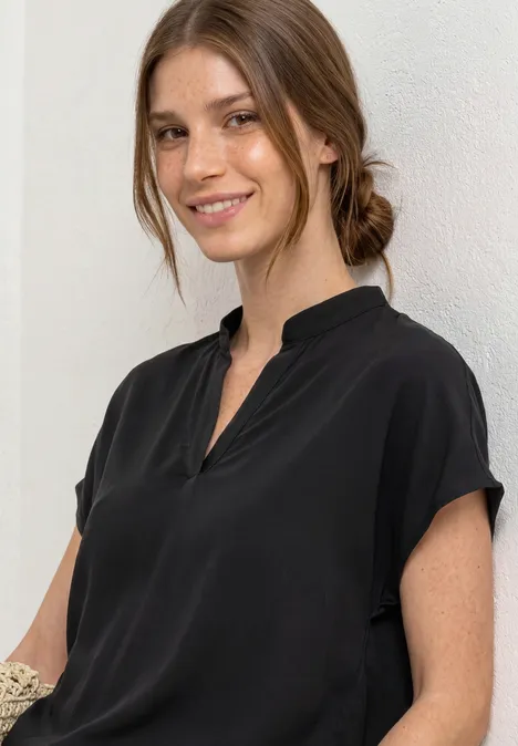 Chemisier à col chemise et ourlet élastiqué Black Femme en chemisier noir à manches courtes, de profil devant un mur blanc.
