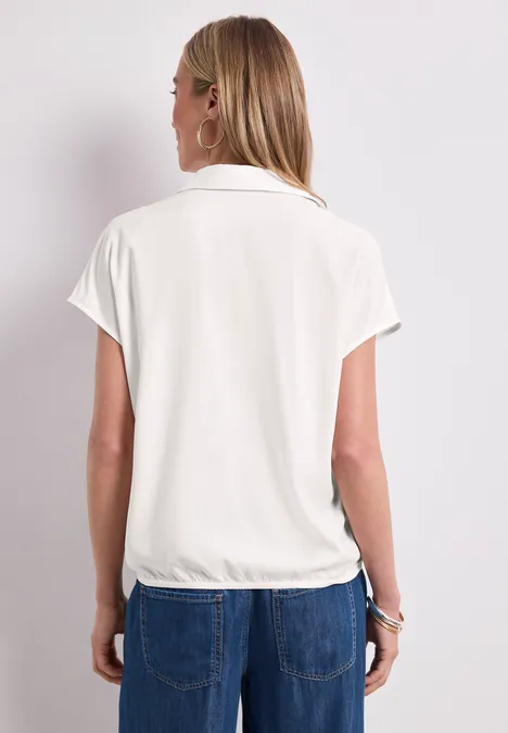 Rückansicht einer Frau in weißer kurzärmeliger Bluse mit Kragen, locker tailliert, dazu blaue Jeans.